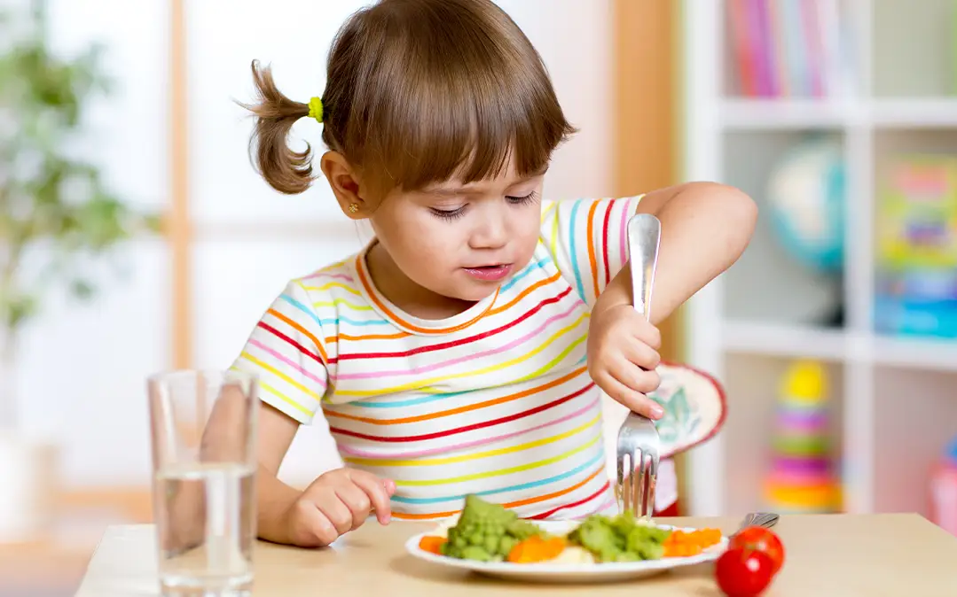 criança pequena, em idade pré-escolar, come prato de legumes, com água à vista 