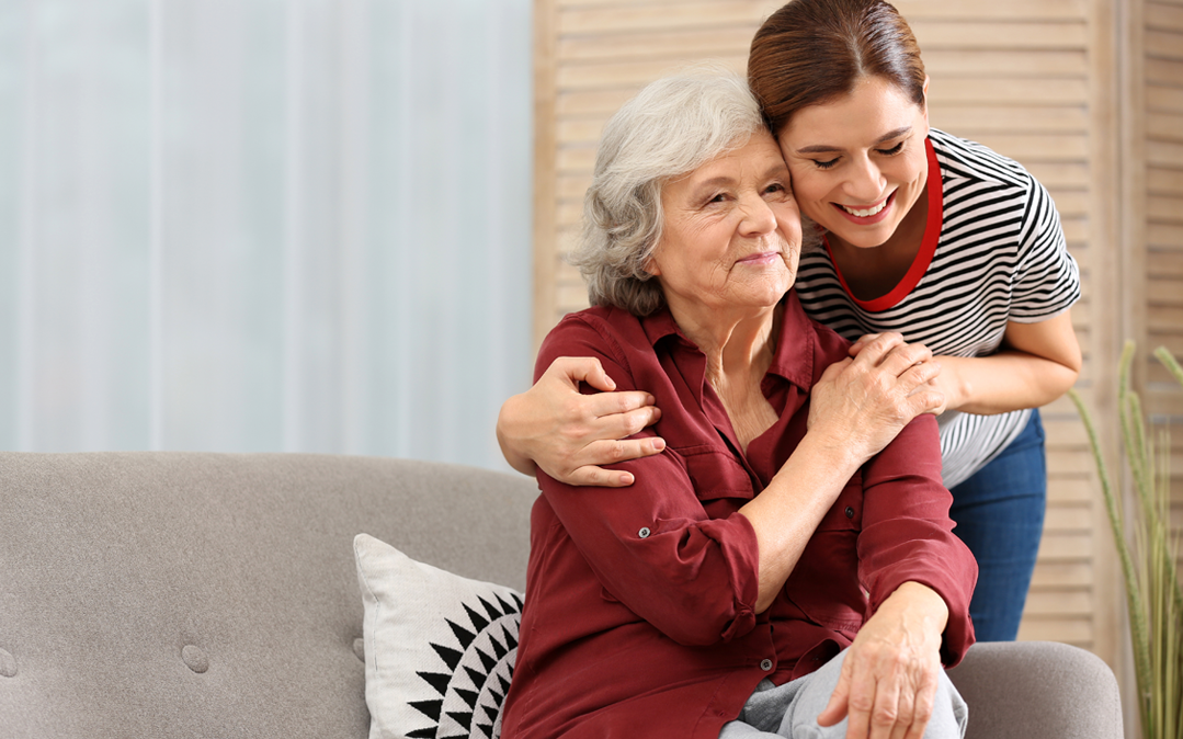 senhora idosa, sentada num sofá, recebe o apoio de outra pessoa, familiar ou cuidador. A  imagem representa a importância do diagnóstico precoce de Alzheimer no bem-estar e qualidade de vida do doente com esta doença.