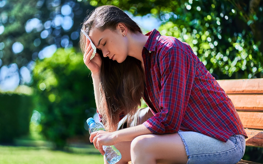 Jovem sentada num jardim, visivelmente cansada pelo calor, limpa o suor da testa