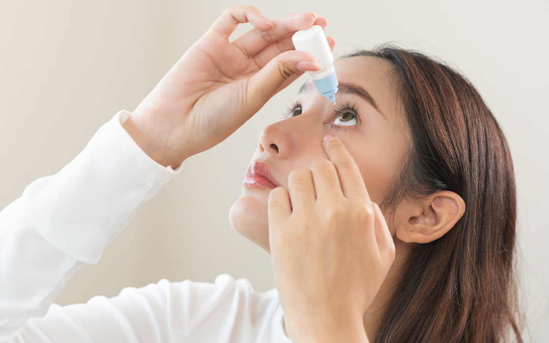 Mulher aplicando colírio no olho direito, com a cabeça inclinada para cima e segurando o frasco com a mão direita, em um ambiente interno iluminado