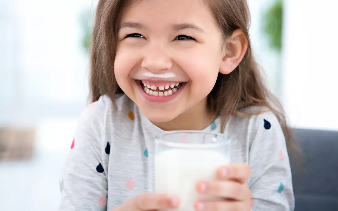 criança pequena com um enorme sorriso, tem um copo de leite na mão e um bigode de leite