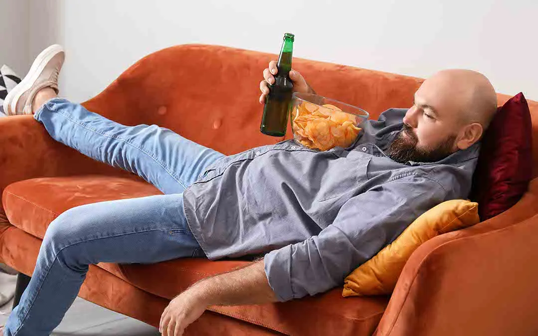 homem deitado ao sofá a comer lanches não saudáveis e a apresentar um comportamento sedentário 